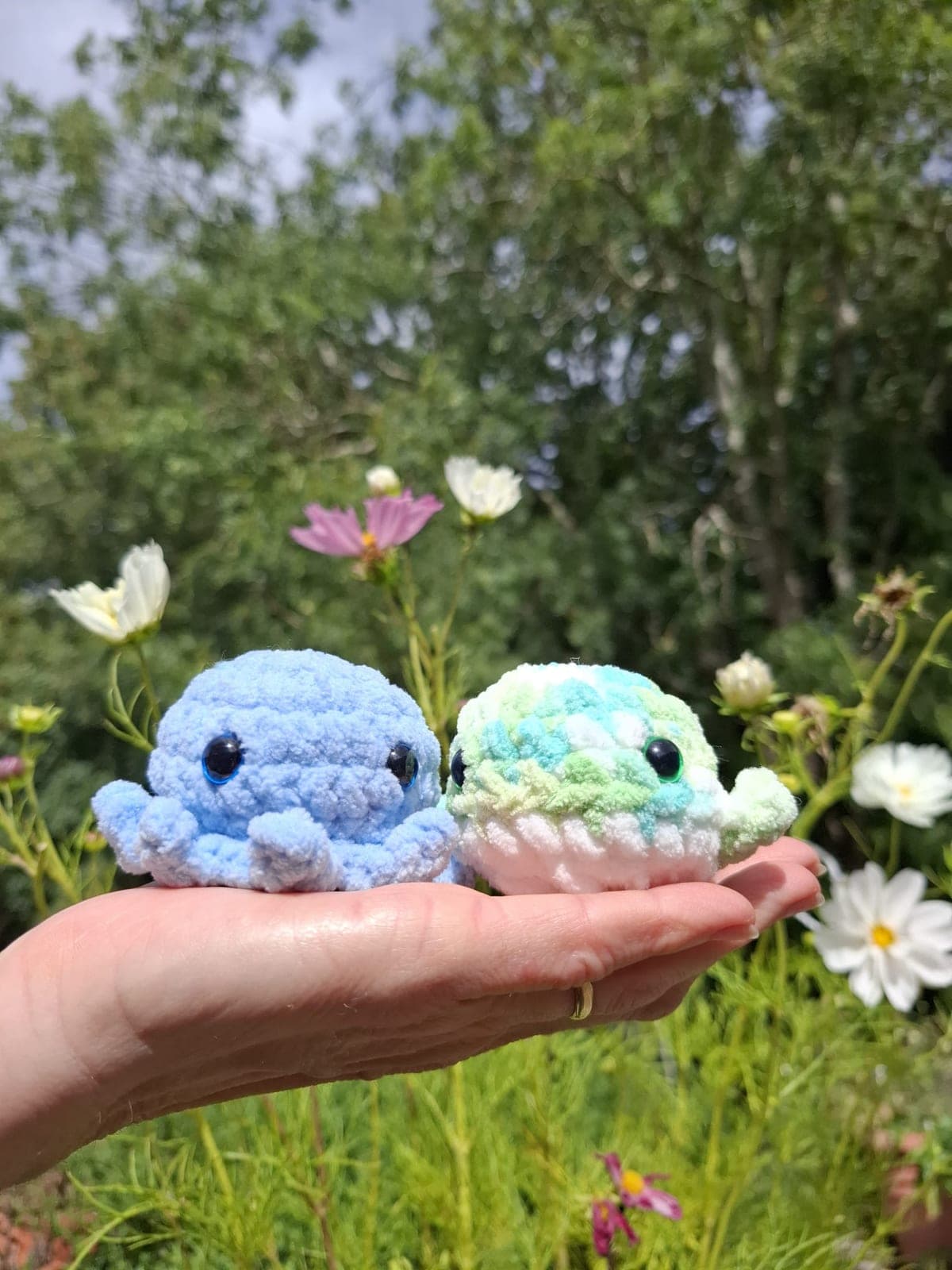 Two small crocheted amigurumi held in a hand among garden flowers